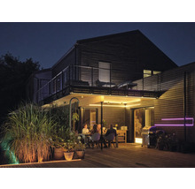 Terrasse en bois éclairée de nuit avec des personnes à table, un barbecue et des bandes LED sous le balcon et au sol.