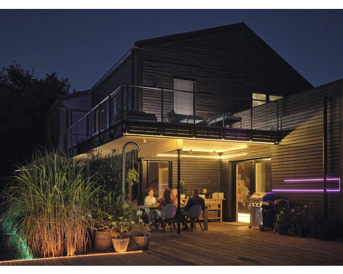 Terrasse en bois éclairée de nuit avec des personnes à table, un barbecue et des bandes LED sous le balcon et au sol.