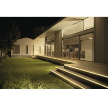 Maison moderne de nuit avec des rubans LED éclairant les marches d'escalier en bois et la terrasse à côté de la pelouse.