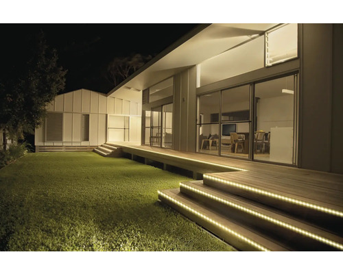 Maison moderne de nuit avec des rubans LED éclairant les marches d'escalier en bois et la terrasse à côté de la pelouse.