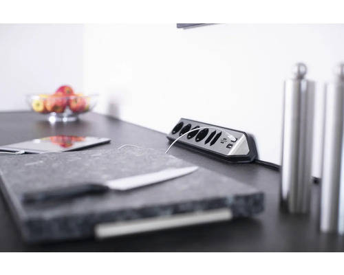 Scène avec une multiprise, une tablette, une planche à découper, un couteau et un saladier de pommes sur un plan de travail.