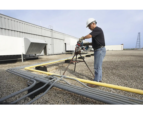 Un homme en vêtements de travail coupe des tuyaux sur un toit avec une scie à tubes.