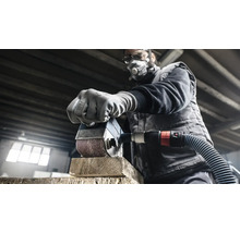 Un homme ponce un morceau de bois avec une ponceuse. Il porte un masque de protection et des lunettes de protection.