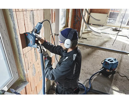 Un artisan utilise une rainureuse à murs sur un mur en briques. Un système d'aspiration de poussière est connecté.