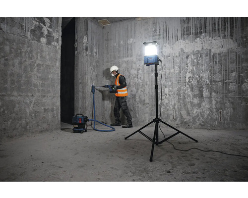 Ein Arbeiter bohrt mit einer Bohrmaschine ein Loch in eine Betonwand, unterstützt von einer Baustellenbeleuchtung und einem Staubsauger.