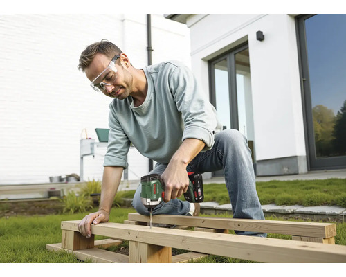 Mann mit Schutzbrille verschraubt Holzbalken mit einem Bosch Akku-Drehschlagschrauber im Garten vor einem Haus.