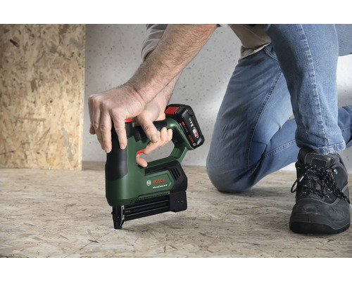 Une personne utilise une agrafeuse sans fil Bosch AdvancedTacker 18 Volt sur un panneau en bois au sol dans une pièce lumineuse.