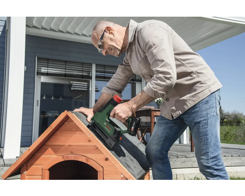 Un homme fixe du feutre bitumé sur le toit d'une niche en bois avec une agrafeuse sans fil Bosch sur une terrasse lumineuse.