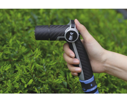 Une main tient un pistolet d'arrosage de la marque For Q devant un buisson vert