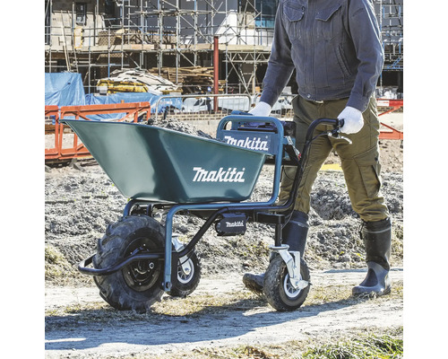 Une personne pousse une brouette à batterie Makita remplie de terre sur un chantier. Appareil bleu à trois roues et Makita Logo.