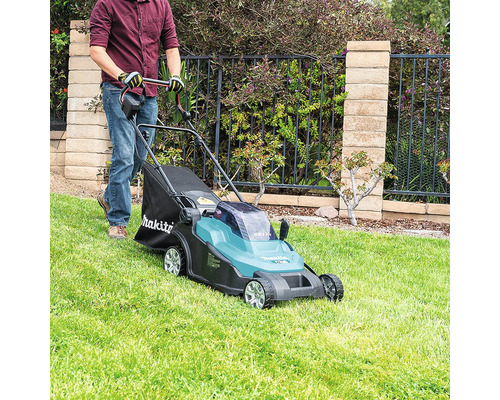 Une personne utilise une tondeuse à gazon sans fil Makita sur une pelouse soignée devant une clôture de jardin avec mur en pierre.