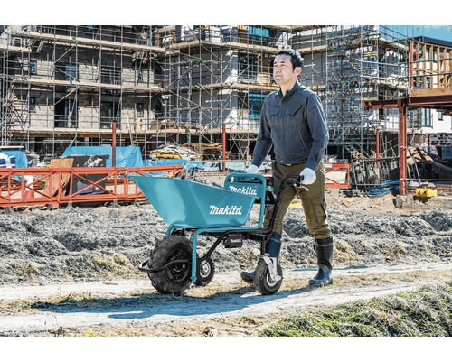 Ouvrier poussant une brouette à batterie Makita sur un chantier de construction avec des échafaudages.
