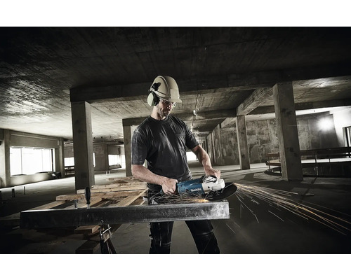 Handwerker mit Schutzhelm und Gehörschutz bearbeitet einen Metallträger mit einem Makita Winkelschleifer in einem Rohbau aus Beton.