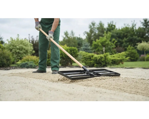 Eine Person ebnet Sand mit einer Einebnungsharke im Garten.