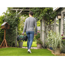 Femme marchant avec un robot de tonte dans un jardin avec une cabane à oiseaux et des plantes