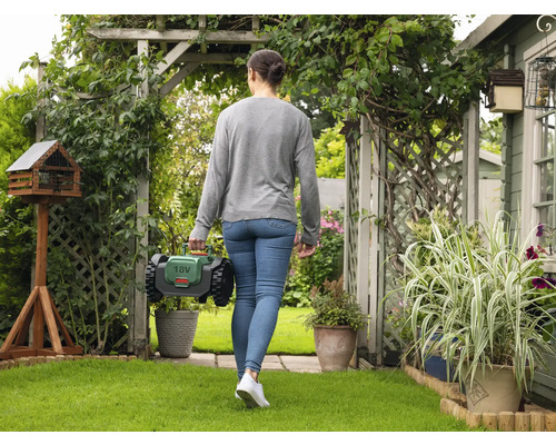 Femme marchant avec un robot de tonte dans un jardin avec une cabane à oiseaux et des plantes