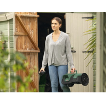 Femme portant un robot tondeuse devant une remise de jardin.