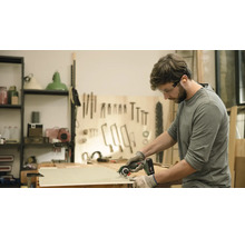 Ein Mann schneidet mit einer Handkreissäge ein Holzbrett. Er trägt dabei eine Schutzbrille und Handschuhe.