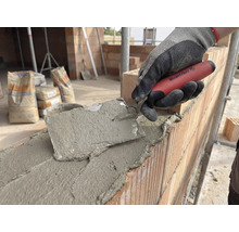 Maçon travaillant avec une truelle sur un mur en briques, sacs de ciment en arrière-plan.