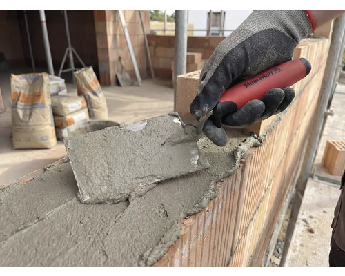 Maçon travaillant avec une truelle sur un mur en briques, sacs de ciment en arrière-plan.