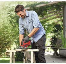 Un homme scie une planche de bois à l'extérieur avec une scie sauteuse. Il porte des lunettes de protection, une blouse de travail et des gants de travail.