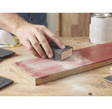 Hand schleift ein lackiertes Holzbrett mit einem Schleifvlies auf einem Schleifklotz. Schleifstaub ist auf dem Holz sichtbar.