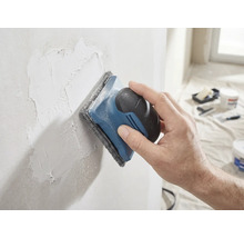 Une main ponce un mur enduit avec une ponceuse à main et un tampon abrasif.