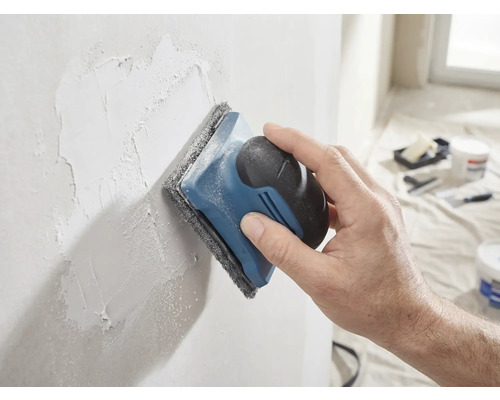 Une main ponce un mur enduit avec une ponceuse à main et un tampon abrasif.