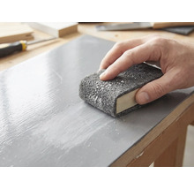 Une main ponce une surface en bois laquée avec un non-tissé abrasif enroulé autour d'un bloc à poncer.