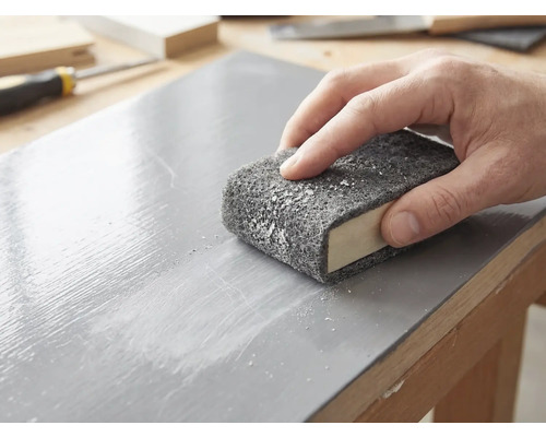 Une main ponce une surface en bois laquée avec un non-tissé abrasif enroulé autour d'un bloc à poncer.