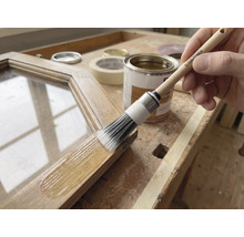 Application d'une lasure pour bois sur un cadre en bois avec un pinceau rond Rautner sur un établi lumineux avec pot de peinture et ruban adhésif.