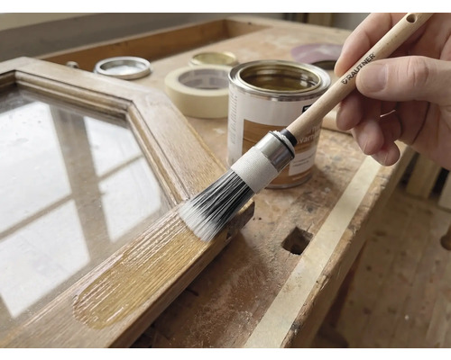 Application d'une lasure pour bois sur un cadre en bois avec un pinceau rond Rautner sur un établi lumineux avec pot de peinture et ruban adhésif.