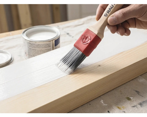 Ein Pinsel mit Holzgriff und roter Manschette trägt weiße Farbe auf einen Holzbalken auf. Im Hintergrund steht eine geöffnete Farbdose.