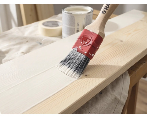 Ein Pinsel mit Holzstiel trägt weiße Farbe auf ein Holzbrett auf, im Hintergrund sind ein Farbeimer und Klebeband zu sehen.