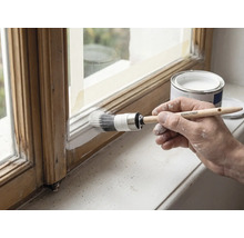 Une main peint en blanc un cadre de fenêtre en bois avec un pinceau rond Rautner, à côté d'un pot de peinture ouvert.