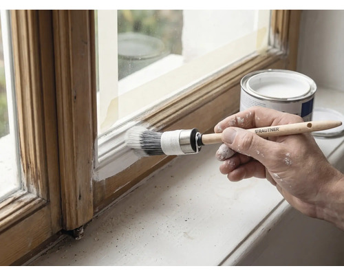 Une main peint en blanc un cadre de fenêtre en bois avec un pinceau rond Rautner, à côté d'un pot de peinture ouvert.