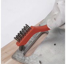 Main avec gant de travail nettoyant une surface métallique avec une petite brosse métallique à manche orange.