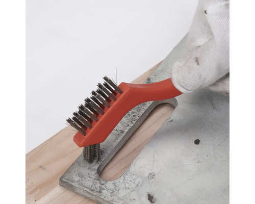 Main avec gant de travail nettoyant une surface métallique avec une petite brosse métallique à manche orange.