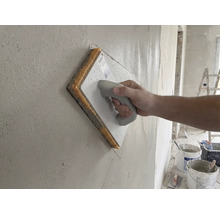 Hand déi e Schwammbriet op enger frësch verputzter Mauer benotzt.