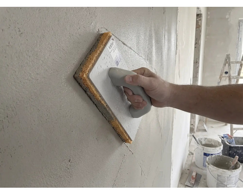 Hand déi e Schwammbriet op enger frësch verputzter Mauer benotzt.