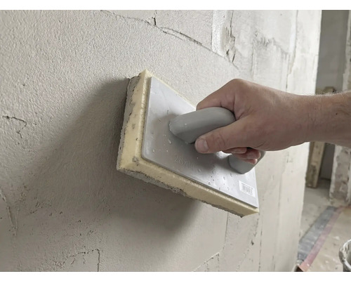 Main utilisant une taloche éponge de 28 par 14 centimètres pour lisser l'enduit frais sur un mur.