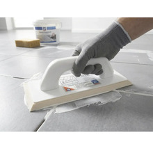 Une personne avec un gant de protection applique du mortier de jointoiement blanc sur des carreaux gris avec une taloche à joints époxy.