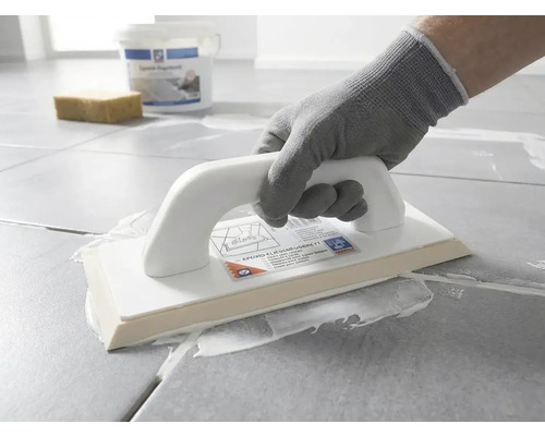 Une personne avec un gant de protection applique du mortier de jointoiement blanc sur des carreaux gris avec une taloche à joints époxy.