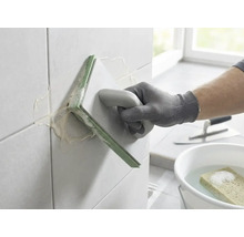 Eine Person verfugt Wandfliesen mit einem Fugengummi. Im Hintergrund befinden sich ein Eimer mit Wasser und Schwamm sowie eine Glättkelle.