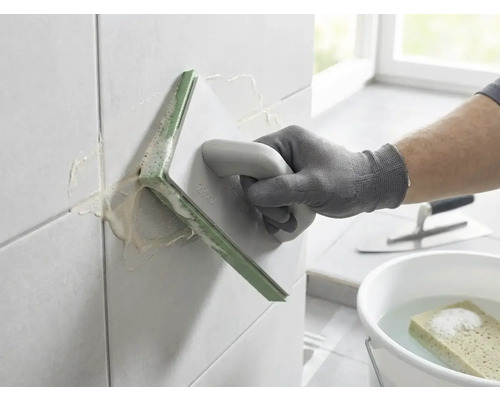 Eine Person verfugt Wandfliesen mit einem Fugengummi. Im Hintergrund befinden sich ein Eimer mit Wasser und Schwamm sowie eine Glättkelle.