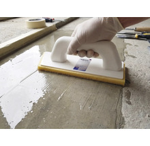 Une personne utilise une taloche à éponge avec poignée blanche et éponge jaune sur une surface en ciment humide.