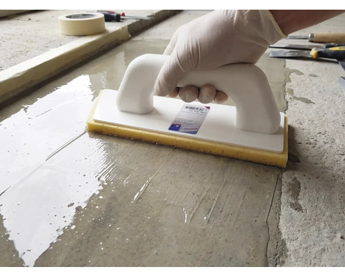 Une personne utilise une taloche à éponge avec poignée blanche et éponge jaune sur une surface en ciment humide.