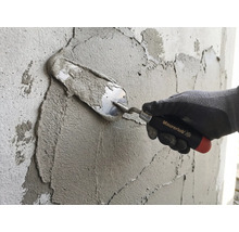 Une main avec un gant de travail applique du mortier sur un mur à l'aide d'une truelle Maurerlob.