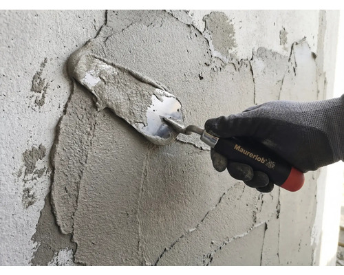 Une main avec un gant de travail applique du mortier sur un mur à l'aide d'une truelle Maurerlob.