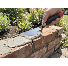 Main avec truelle Maurerlob appliquant du mortier sur un mur en briques dans un jardin.